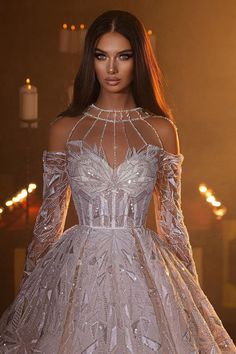 a woman in a white dress with long sleeves and beading on the shoulders, standing next to candles