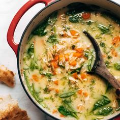 a red pot filled with soup and bread on top of a white tablecloth next to a wooden spoon