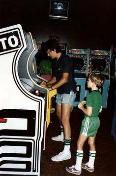 two young boys playing video games in a room with arcade machines and other people standing around