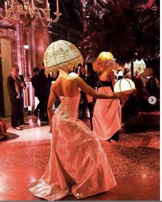 two women in dresses and hats are walking down the hall with an umbrella over their head