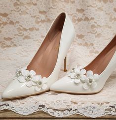 a pair of white high heel shoes with flowers on the side and lace around the toe