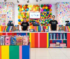 two men standing in front of a rainbow colored candy shop with balloons on the wall