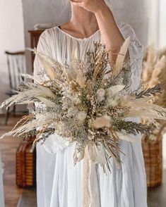 a woman in a white dress holding a bouquet of dried flowers and grasses with her hands to her face