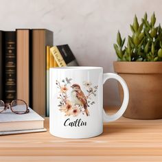 a coffee mug with a bird on it sitting next to some books and a potted plant