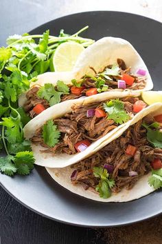 two shredded beef tacos on a plate with cilantro and lime wedges