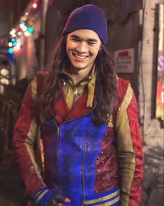 a man with long hair wearing a blue and red jacket, smiling at the camera