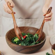 a person holding two wooden utensils over a bowl filled with green vegetables and broccoli