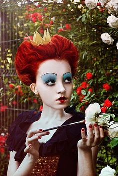 a woman with red hair and makeup holding a flower in front of her face while wearing a crown