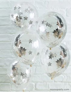 five clear balloons with silver stars and confetti on them, against a white brick wall