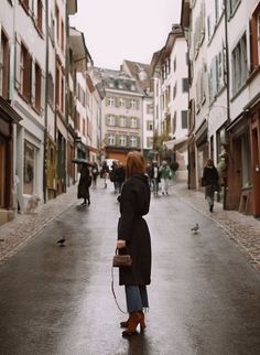 a woman standing in the middle of an alley way with buildings on both sides and people walking down the street