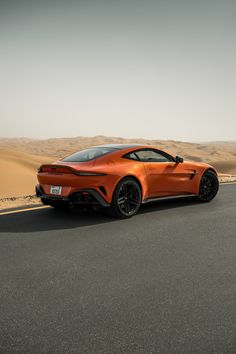 an orange sports car is driving down the road in the middle of the desert with sand dunes behind it