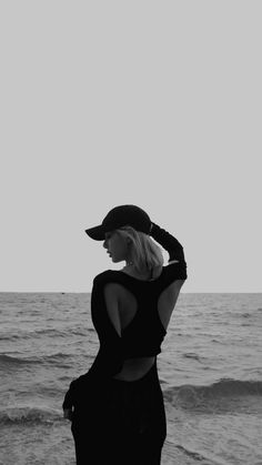 a woman standing on top of a beach next to the ocean wearing a baseball cap