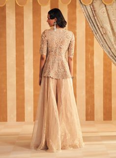 Shine in the champagne net peplum with tissue flared sharara. The full embroidery on the net fabric adds a luxurious touch, complemented by the soft, flowing tissue sharara. This ensemble is perfect for formal occasions, combining opulent detailing with a contemporary silhouette for a truly elegant look. Glamorous Floor-length Sharara With Resham Embroidery, Beige Floor-length Party Sets, Glamorous Sharara With Resham Embroidery For Reception, Glamorous Resham Embroidery Sharara For Reception, Glamorous Resham Embroidered Sharara For Reception, Gold Palazzo Set For Wedding, Gold Palazzo Set For Wedding Party, Glamorous Organza Sharara For Festive Occasions, Elegant Peplum Lehenga For Reception