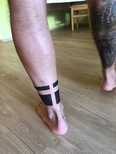 a man's foot with a cross tattoo on his left leg, and a wooden floor in the background