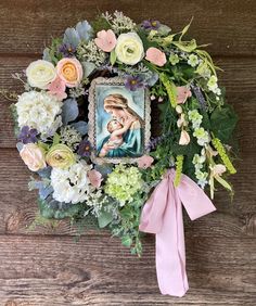 a wreath with an image of the virgin mary surrounded by flowers and greenery on a wooden surface