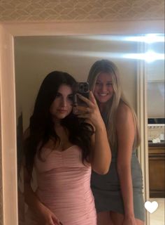 two beautiful young women standing in front of a mirror taking a selfie with their cell phone