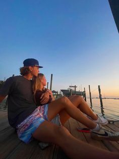 a man and woman are sitting on a dock looking at the water as the sun sets