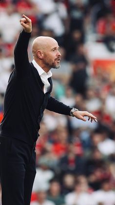 a bald man in a black suit and white shirt gestures to his left while standing on the sidelines