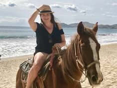 a woman riding on the back of a brown horse down a sandy beach next to the ocean