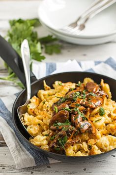 a skillet filled with pasta and mushrooms on top of a wooden table next to silverware