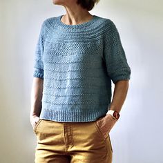 a woman standing in front of a white wall wearing a blue sweater and gold pants