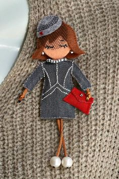 the doll is wearing a gray dress and holding a red purse in her hand while standing next to a knitted sweater