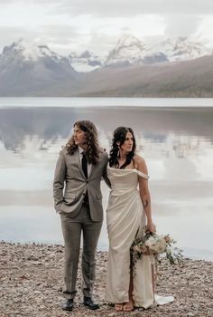 a man and woman standing next to each other on a beach near the water with mountains in the background