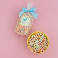 a small yellow bowl filled with candy corn next to a bag of jelly beans on a pink background