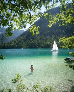 a person standing in the water with a sailboat and mountains in the back ground