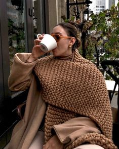 a woman sitting on a bench drinking from a coffee cup while wrapped in a blanket