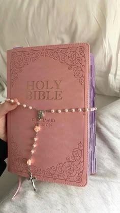 a pink bible with beads and a cross on the cover is held up by a woman's hand