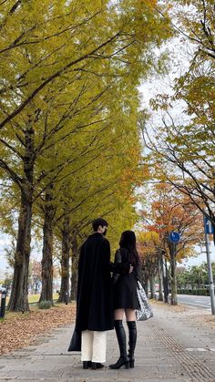 two people dressed in black and white are walking down the street with trees lining both sides