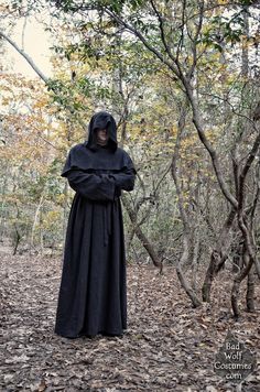 a man in a black hooded costume standing in the woods with his arms folded up