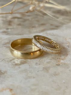 Vintage-inspired wedding bands in yellow gold stacked. Rounded men's band with a brush finish and milgrain texture detailing along the borders. Women's wedding band is an eternity channel set style with baguette diamonds vertically set between a similar milgrain texture detailed border for a matching wedding band set. Simple Elegant Rings Gold, Wedding Rings His, Wedding Bands Men And Women, Wedding Bands His And Hers Gold, Wedding Ring Gold And Silver, Gold Wedding Band Sets His And Hers, Wedding Band Mixed Metals, Simple Wedding Band Set, Roman Numeral Wedding Band