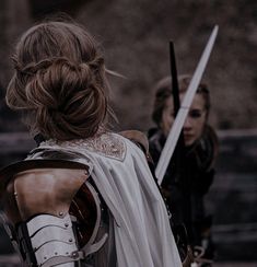 two women dressed in medieval costumes holding swords and looking at each other while standing next to one another