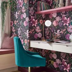 a desk with a blue chair in front of a flowered wallpaper and window