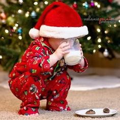 a baby in pajamas and santa hat drinking milk from a bottle next to a christmas tree