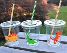 three plastic cups with different colored straws in them on a picnic table near some bushes