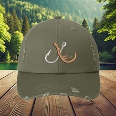 100% Cotton, One size fit the most adults.  Blend your love for fishing and hunting with this stylish olive green distressed baseball cap, featuring a unique embroidered design combining a fishing hook and a deer antler. This cap, made from high-quality cotton, is comfortable, breathable, and perfect for all your outdoor adventures. The distressed look gives it a rugged, well-worn appearance that suits any enthusiast of the great outdoors. The adjustable strap ensures a snug, customizable fit for all head sizes, making it perfect for men and women alike. Whether you're heading out to fish, hunt, hike, or just enjoy the outdoors, this cap is the ideal accessory. It also makes a fantastic gift for hunters, anglers, or anyone with a passion for nature. Features: Material: 100% Cotton Design: Fishers Hat, Outdoorsman Gifts, Hunting Hat, Distressed Baseball Cap, Fishing Hat, Gifts For Hunters, Hunting Trip, Embroidered Hats, Hunting Fishing