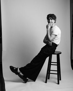 a young man sitting on top of a stool next to a white wall with his legs crossed