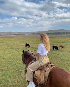 a woman riding on the back of a brown horse in a field full of cows