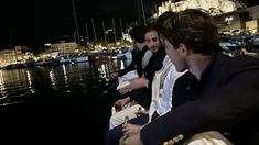 three men sitting next to each other in front of the water at night with boats docked behind them