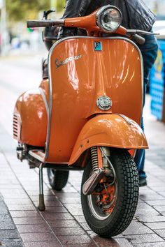 an orange motor scooter parked on the side of a brick road next to a sidewalk