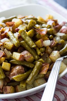 a white bowl filled with cooked green beans and potatoes next to a fork on a table