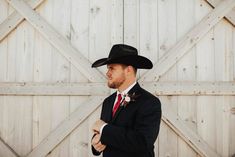 a man in a black suit and cowboy hat standing next to a wooden wall wearing a red tie