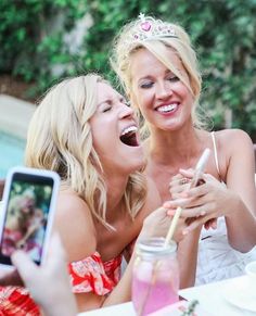 two women laugh as they take pictures with their cell phones at an outdoor table in front of a pool