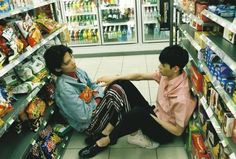 two people sitting on the ground in front of a store filled with drinks and snacks