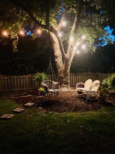 two lawn chairs sitting under a tree with lights strung from it's branches at night