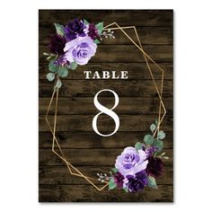 a table number with purple flowers and greenery in the center on a wooden background