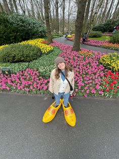 a woman wearing yellow rain boots standing in front of colorful flower garden with trees and bushes